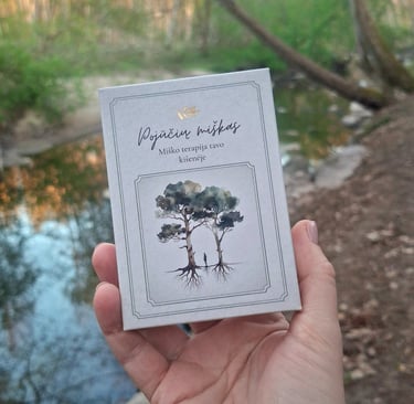 Patyriminės kortelės "Pojūčių miškas", laikomos rankoje, miško ir upelio apsupty.