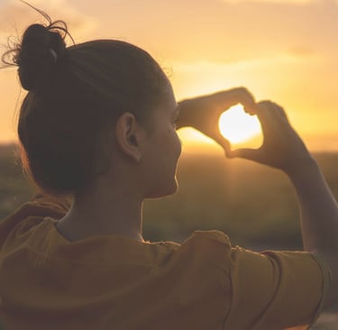 Moteris stovinti saulėlydžio šviesoje pievoje, rodo širdelę ant besileidžiančios saulės.