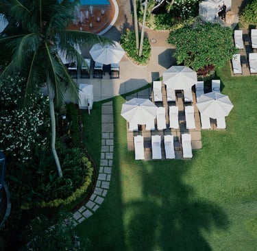 Aerial view of a tropical resort garden with white patio umbrellas and lounge chairs on a lush green lawn.