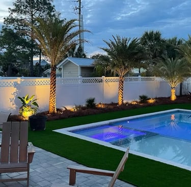 Backyard swimming pool with artificial turf and palm trees illuminated by warm landscape lighting.