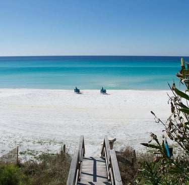 A wooden boardwalk leads to a white sand beach with turquoise ocean water and beach chairs.