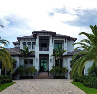 Luxury white two-story coastal mansion with palm trees and a paved driveway under a cloudy sky.