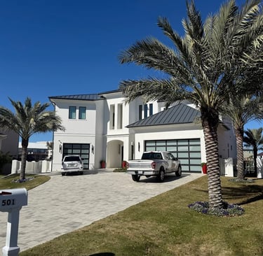 Modern white luxury home with a paved driveway, palm trees, and glass garage doors under a clear blue sky.