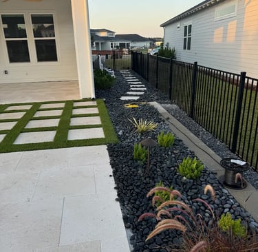Modern backyard landscaping featuring a travertine patio, black river rock, and artificial turf strips.
