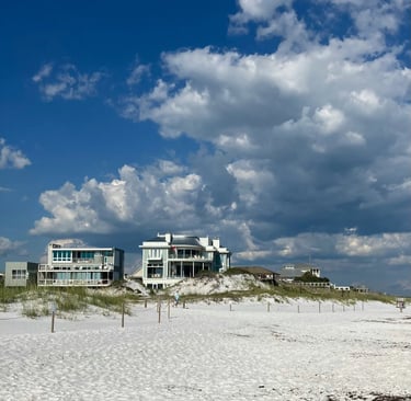 Luxury beachfront vacation rentals on white sand dunes under a blue sky with fluffy clouds.