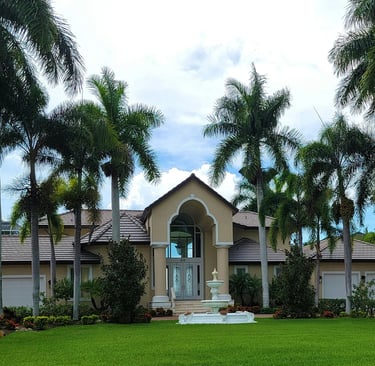 Luxury Florida mansion with tall palm trees, manicured green lawn, and a tiered garden fountain.