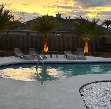 Backyard swimming pool with lounge chairs and lighted palm trees at sunset.