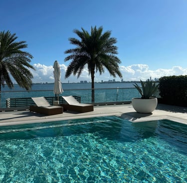 Luxury infinity pool with lounge chairs and palm trees overlooking a tropical bay and city skyline.