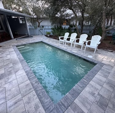 A rectangular backyard plunge pool with stone pavers and white Adirondack chairs in a lush patio setting.