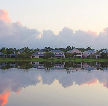 Luxury waterfront homes reflected in a calm lake under a pastel pink sunset sky with fluffy clouds.