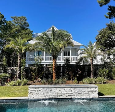 Luxury backyard swimming pool with a stone waterfall feature, palm trees, and a white beach house.