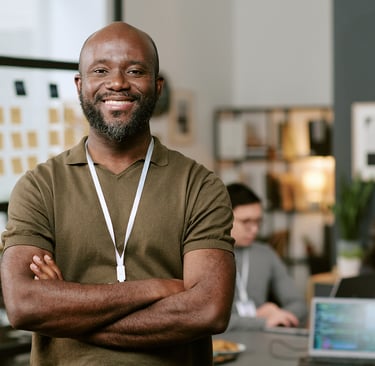 A smiling Black professional male standing with arms crossed in a modern open-plan office.