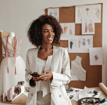 A smiling fashion designer holding a smartphone in her studio with sketches and a mannequin.