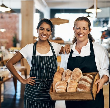 Two smiling female bakers holding a wooden crate of fresh artisanal bread loaves in a bakery.