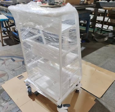 White metal rolling storage cart with shelves wrapped in plastic film for shipping in a factory.