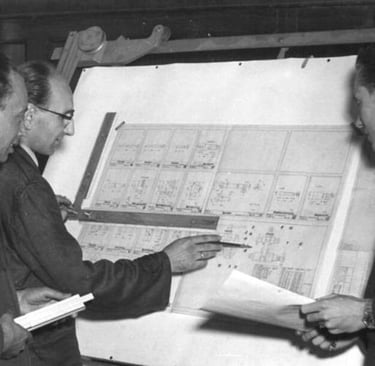 Vintage black and white photo of three engineers reviewing technical blueprints on a drafting board.