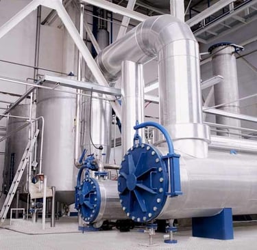 Industrial factory interior featuring stainless steel storage tanks and large insulated piping systems.