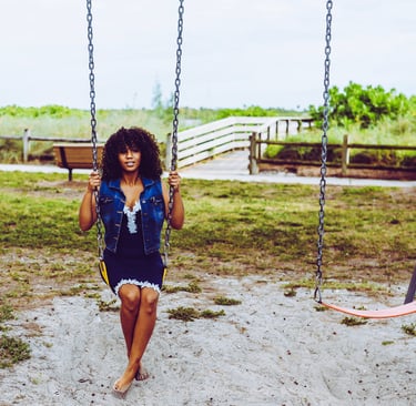 A young woman with curly hair sitting on a playground swing set in an outdoor park.