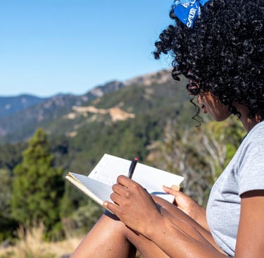 A woman with curly black hair writes in her journal while sitting outdoors with a mountain view.