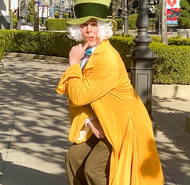 A Mad Hatter inspired performer with a large green top hat and yellow coat poses outdoors.