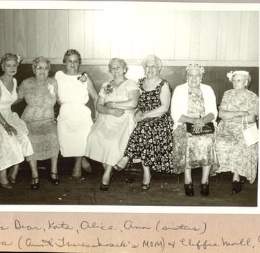 Vintage black and white photo of seven women in floral dresses sitting together at a formal gathering.