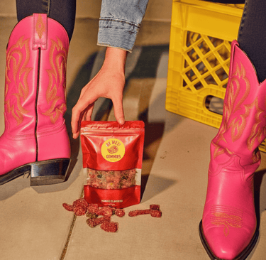 A person wearing vibrant pink western cowboy boots holds a bag of mango flavored gummies.