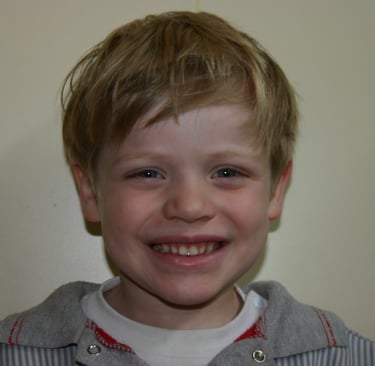 Smiling young boy with messy blonde hair posing for a portrait against a plain wall.