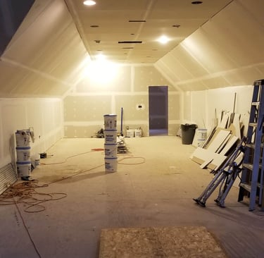 Indoor construction site showing a room with new drywall installation, ceiling lights, and joint compound buckets.