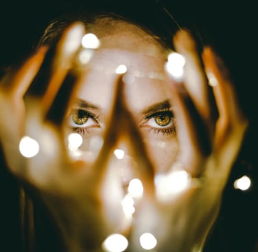 A woman with green eyes peeks through bokeh string lights held in her hands against a dark background.