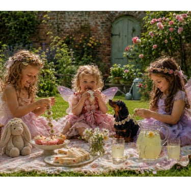 Three little girls dressed as fairies enjoy a picnic with a dachshund wearing a daisy chain & a bunny toy in a walled garden.