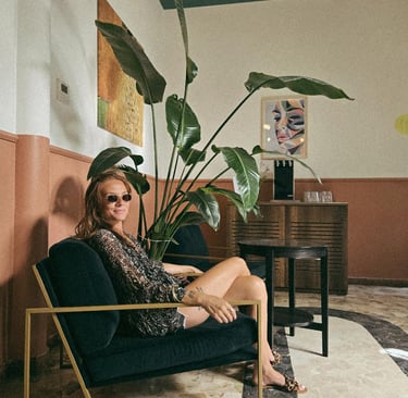 A stylish woman in sunglasses sits in a velvet armchair next to a large bird of paradise plant in a modern cafe.
