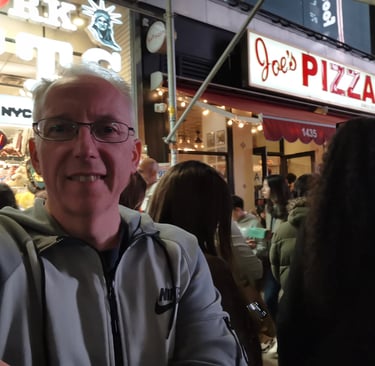 A man in a Nike jacket takes a selfie outside Joe's Pizza in New York City at night.