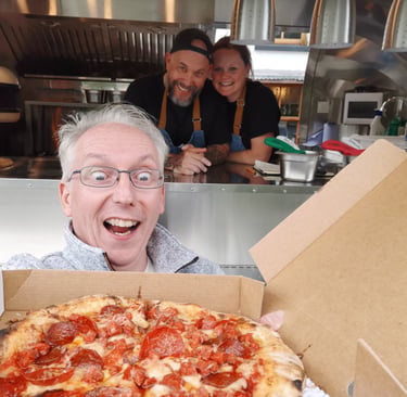 A happy man holds a fresh pepperoni pizza in front of a food truck with two smiling chefs.