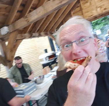 A man wearing glasses eating a slice of wood-fired pizza at an outdoor patio party.