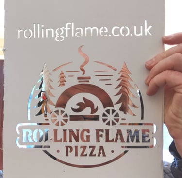 A person holding a laser-cut stencil of the Rolling Flame Pizza logo featuring an outdoor oven and pine trees.