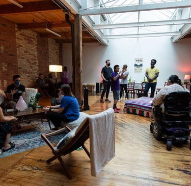 Gallery view of a living room with a bed, poufs, carpet, and about 20 people in the space.