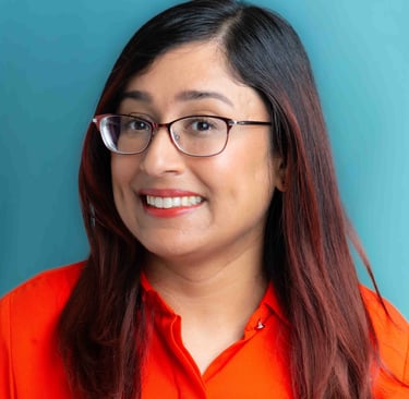 A woman with long, reddish-brown hair and glasses smiles warmly in a bright orange top against a teal background.