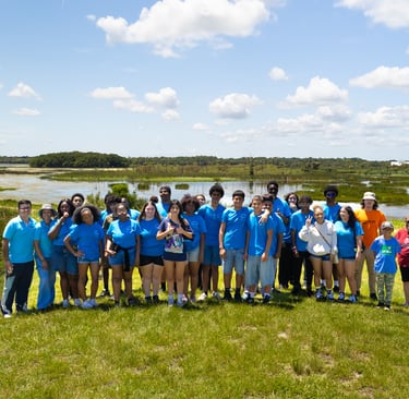 A diverse group of students in blue shirts posing for a photo at a scenic Florida nature preserve wetland.