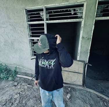 Man in black hoodie and corduroy cap standing outside a rustic, abandoned concrete building.