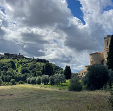 Medieval Abbey of Sant'Antimo in Tuscany surrounded by rolling hills and olive trees under a cloudy blue sky.