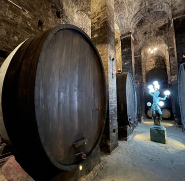 Historic stone wine cellar with large oak barrels and a lit statue under vaulted ceilings.