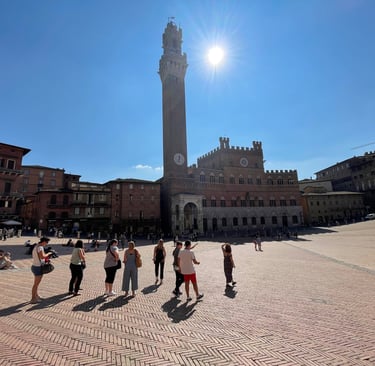 Tourists standing in Piazza del Campo under a bright sun with the Torre del Mangia in Siena, Italy.
