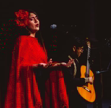 A female singer in a red shawl and flower hair accessory performs with a live acoustic guitarist.