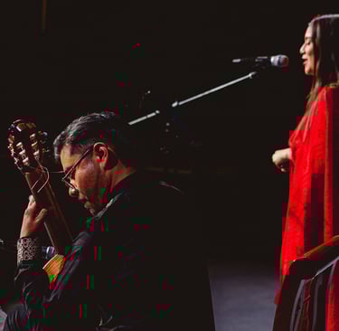 A male guitarist plays acoustic guitar while a female singer in a red dress performs at a live concert.