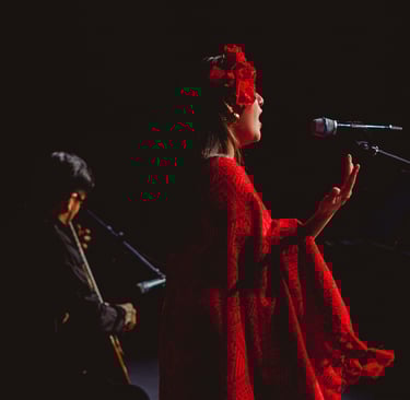 A woman in a red dress with flower hairpiece singing into a microphone during a live musical performance.