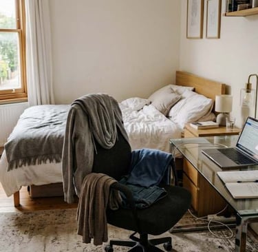 European bedroom with a chair covered in clothes, a desk with a laptop and mug, and a slightly unmade bed