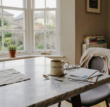 A kitchen table with a mug, scattered post, and a chair with a jumper in soft morning light