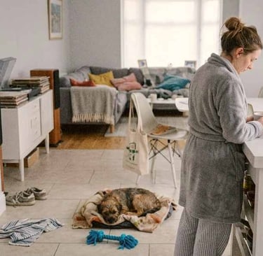Woman in a dressing gown standing in a softly lit European home with a small dog resting nearby and everyday clutter around