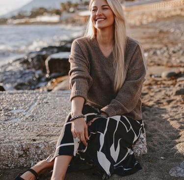 Smiling blonde woman in a brown v-neck sweater and patterned skirt sitting on a beach at sunset. 