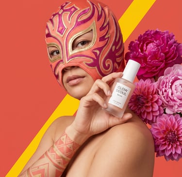 A person in a lucha libre mask holds Glow Seoul facial serum next to pink dahlia flowers.